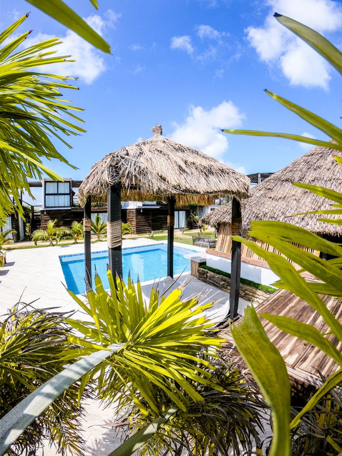 A tranquil resort scene featuring nipa huts by a clear swimming pool under a vibrant blue sky.