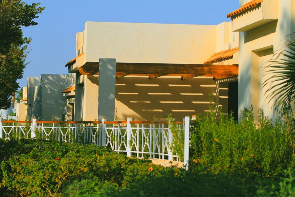 Stylish villa exterior with shaded pergola and lush garden in bright sunlight.